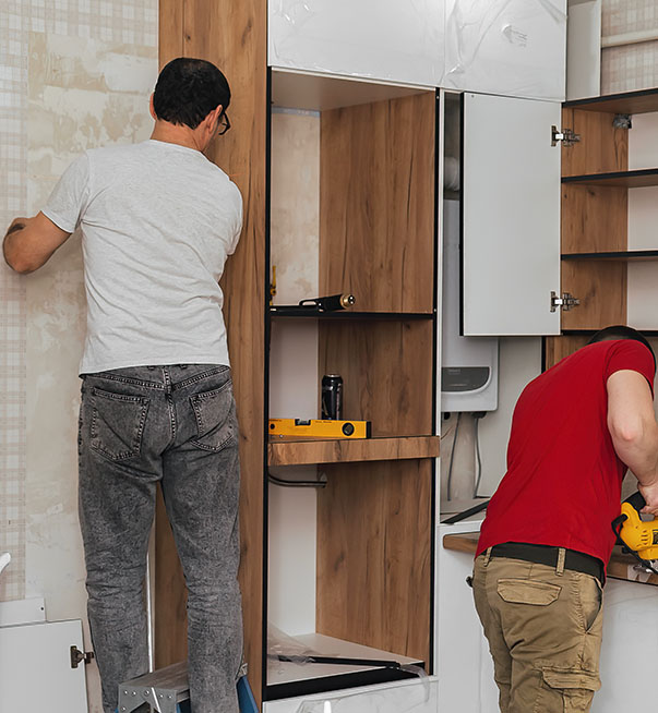 Two workers are installing wooden cabinetry in a kitchen, using tools like a level and a drill, with wallpaper visible in the background.