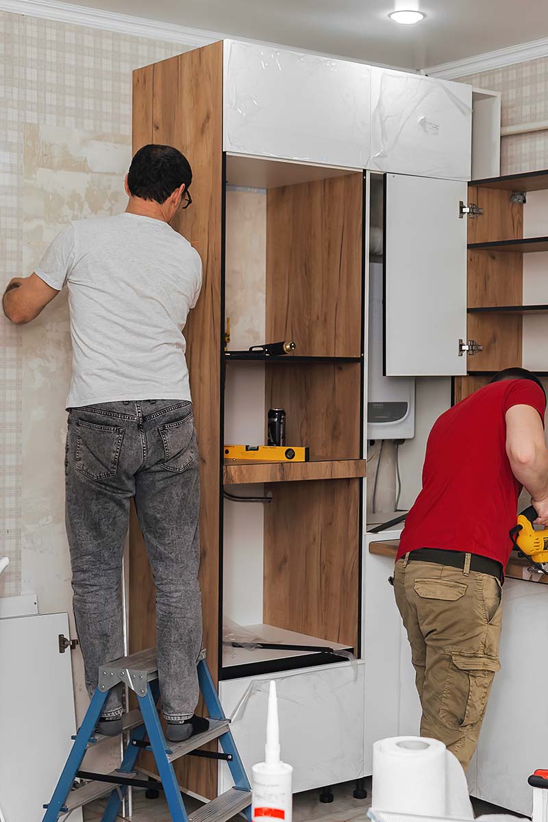 Cabinets being installed around plumbing and electrical wiring in a kitchen.