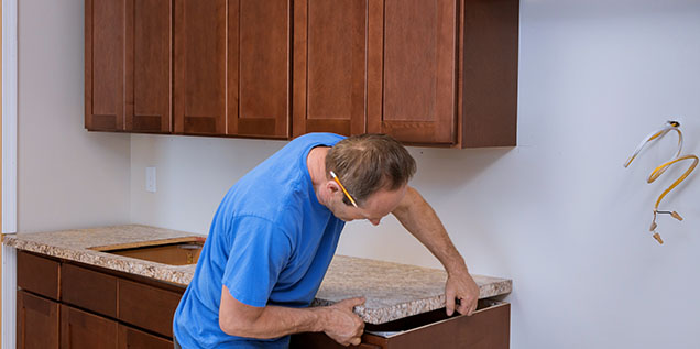 A man in a blue shirt installs a countertop onto brown kitchen cabinets, concentrating on aligning the pieces correctly.